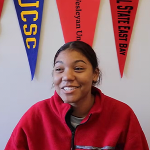 Student smiling at camera, with a variety of college banners behind her