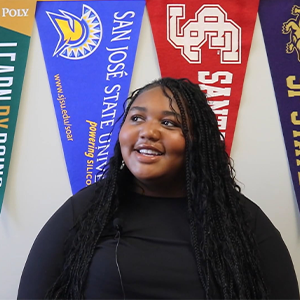 Student smiling at camera, with a variety of college banners in the background