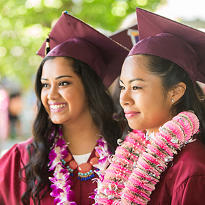 Two students standing outside, wearing graduation caps, graduation gowns, and leis